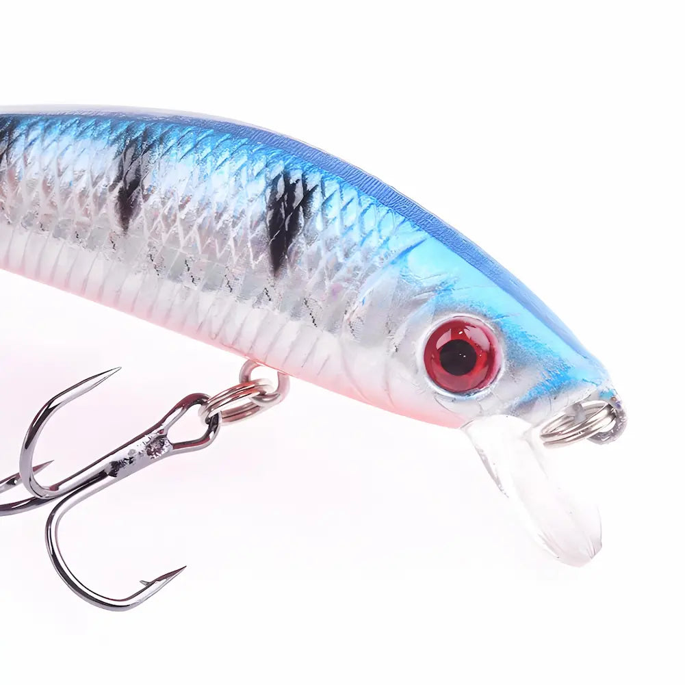 A imagem mostra um close-up de uma isca de pesca artificial com formato de peixe. A isca tem corpo prateado com listras pretas e azul na parte superior, olhos vermelhos grandes e uma pequena pala transparente na frente. Possui um anzol triplo de cor escura preso na parte inferior. O fundo é branco, destacando as cores e detalhes da isca.